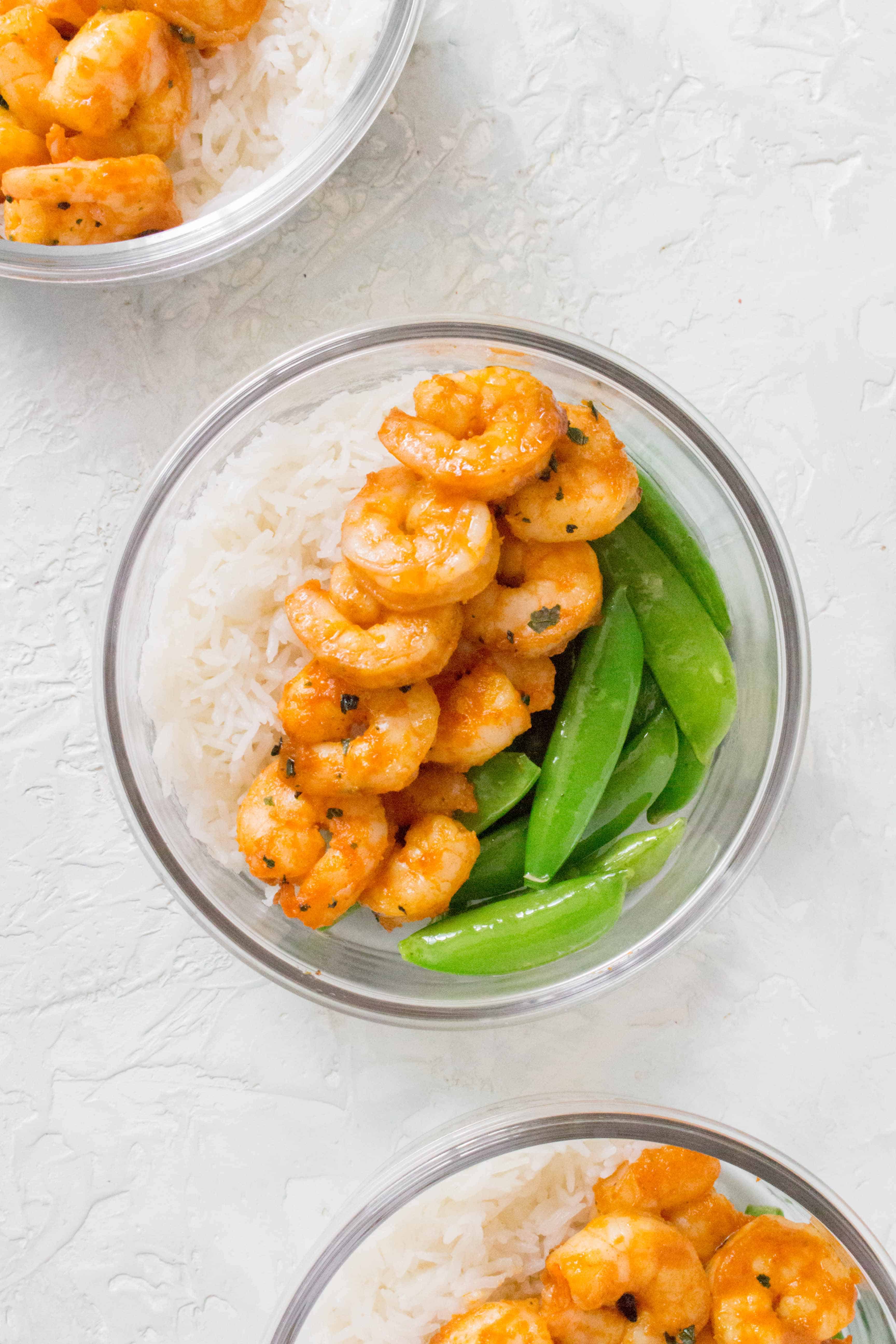 Close up of a glass meal prep container with rice, sriracha lime shrimp, and snap peas.