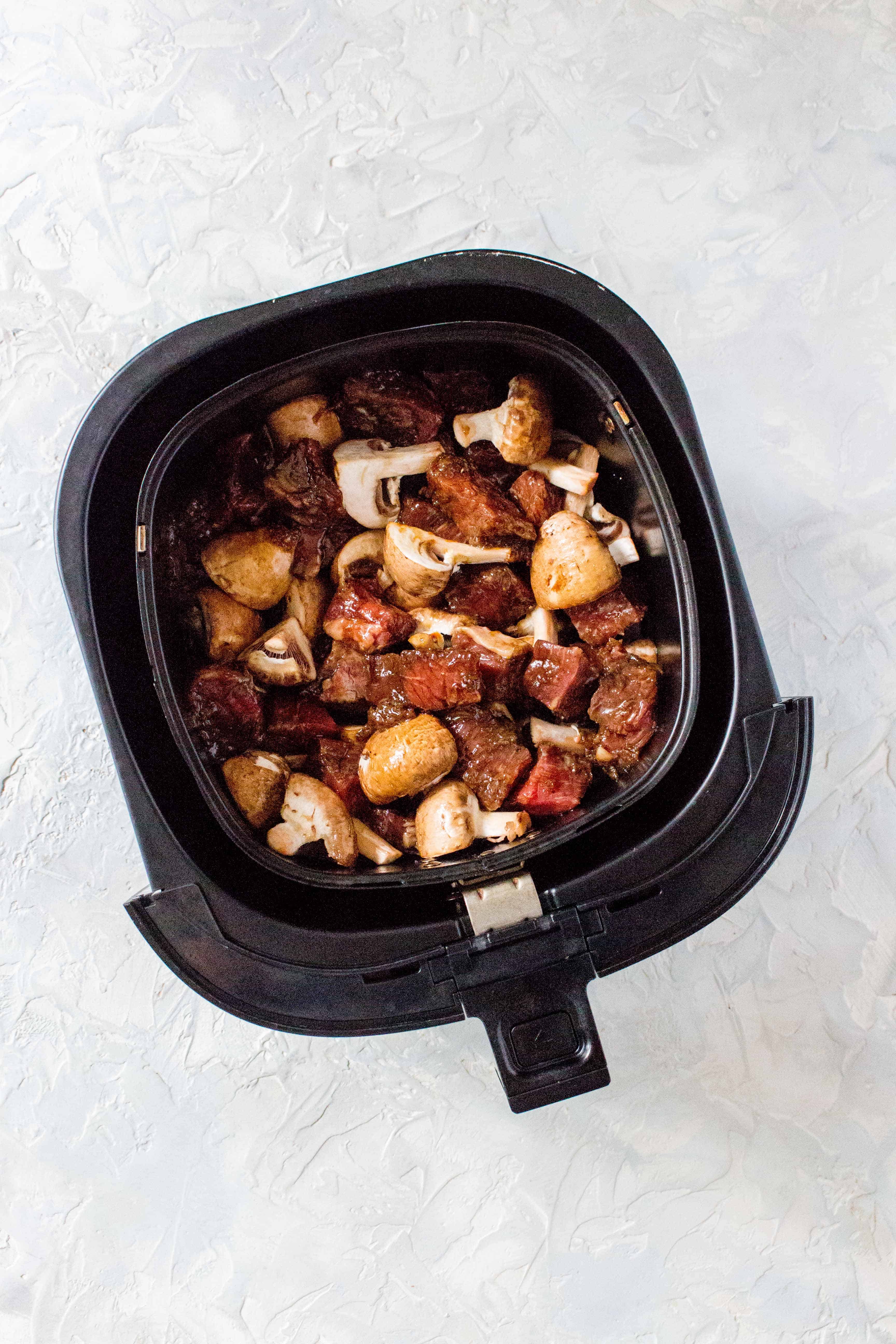 Overhead view of an air fryer basket with marinaded beef and mushrooms.