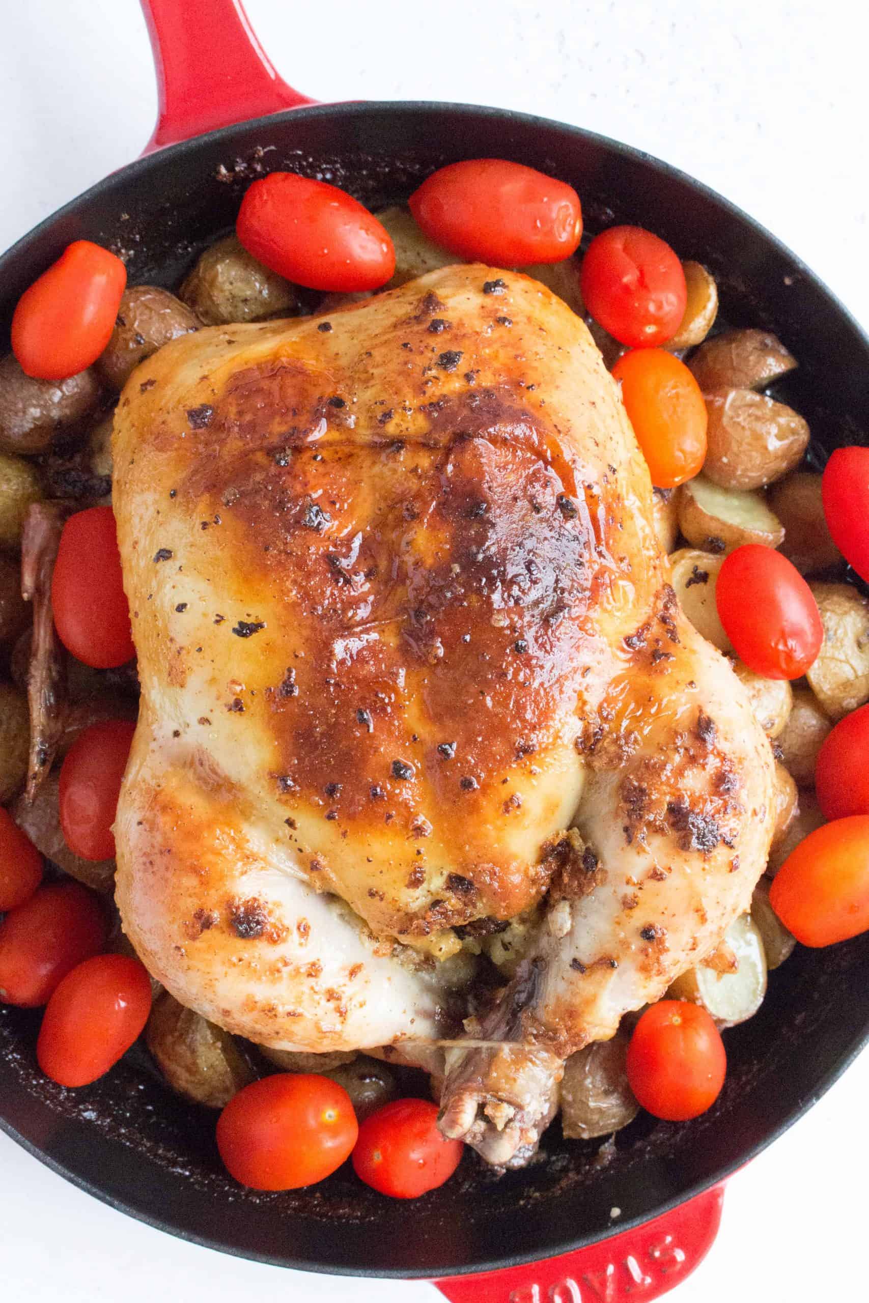 Whole Roast Chicken Inside A Red Cast Iron Skillet Surrounded By Baby Potatoes And Tomatoes