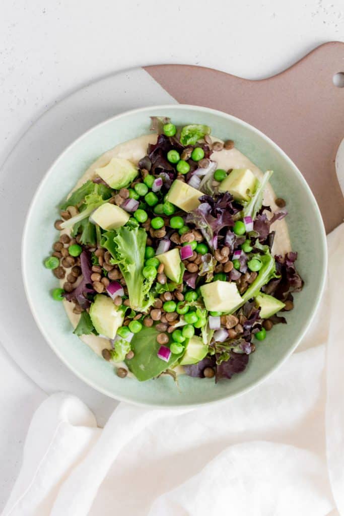 green plate with hummus and mixed greens with green lentils, red onions, avocados, and peas.