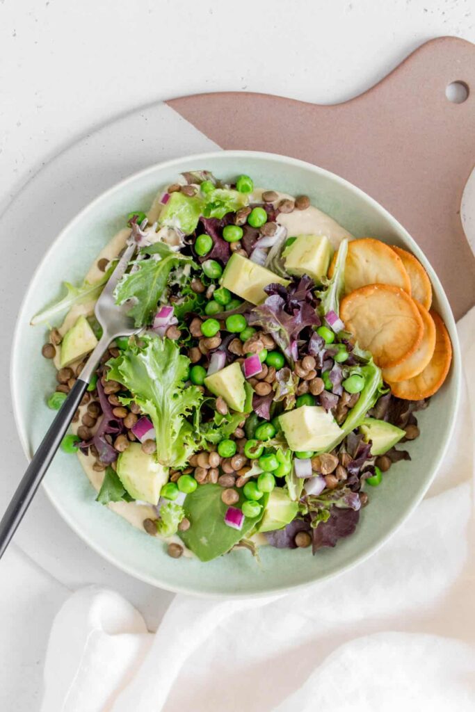 green plate with hummus and mixed greens with green lentils, red onions, avocados, and peas with a fork.