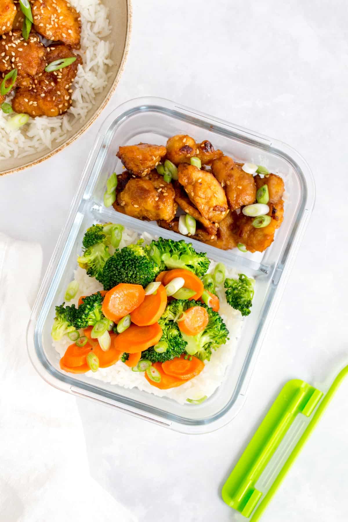Bento box of sesame chicken in a glass meal prep container with rice and vegetables.