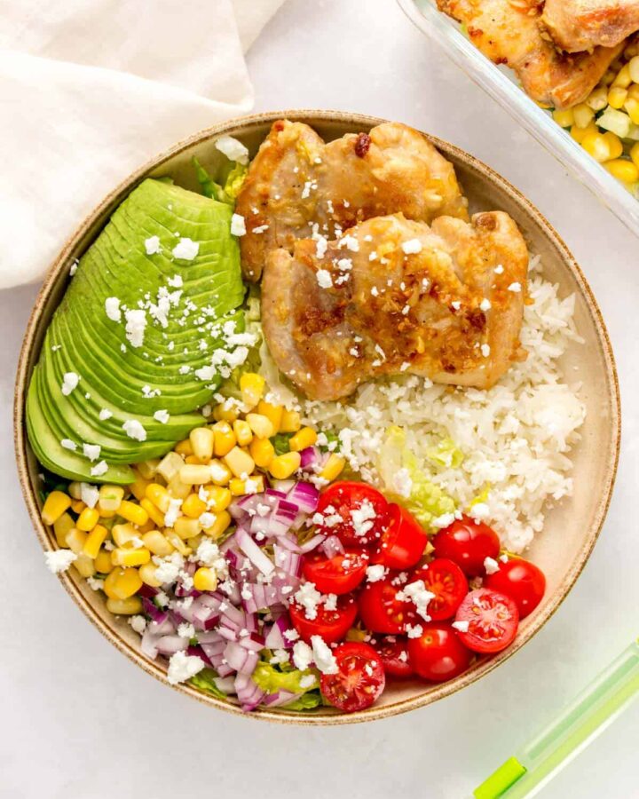 A plate with rice, lettuce, tomatoes, red onion, corn, avocado, and garlic butter chicken thighs.