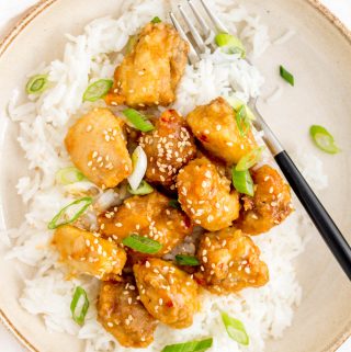 A plate of rice with air fryer orange chicken on top with a fork in the plate. Garnished with sesame seeds and green onion.