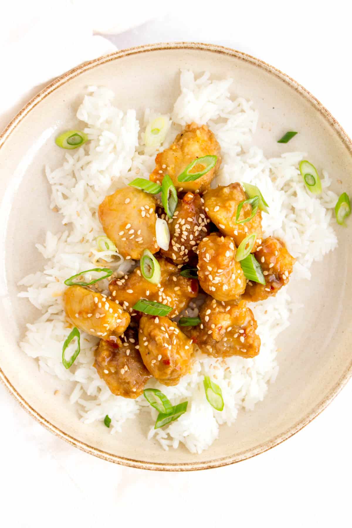 A plate of rice with air fryer orange chicken on top. Garnished with sesame seeds and green onion.