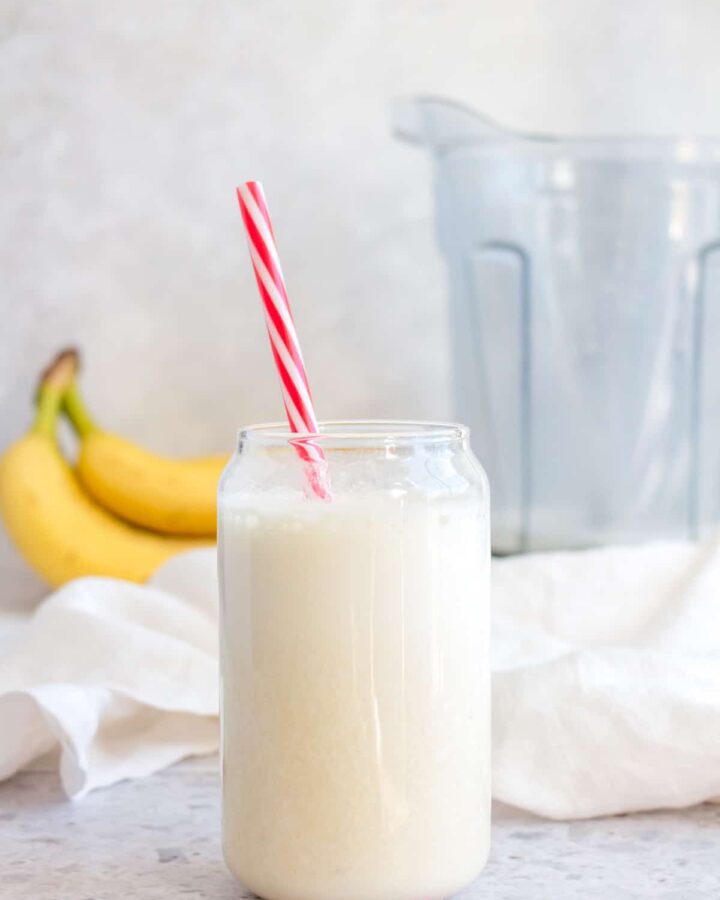 Banana milk in a cup with a red and white straw with bananas and a vitamix in the background.