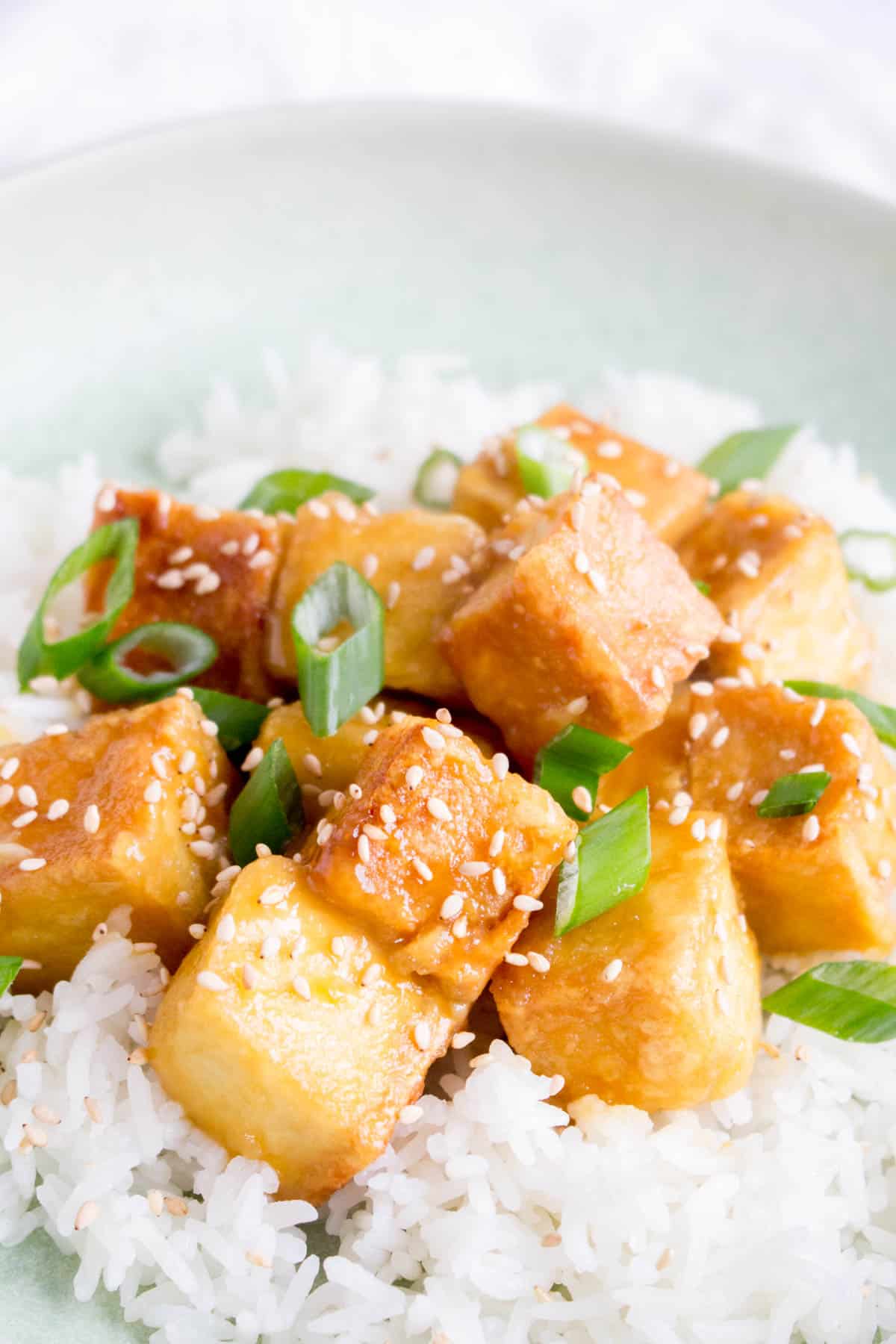 Close up of orange tofu over rice with green onions and sesame seeds.