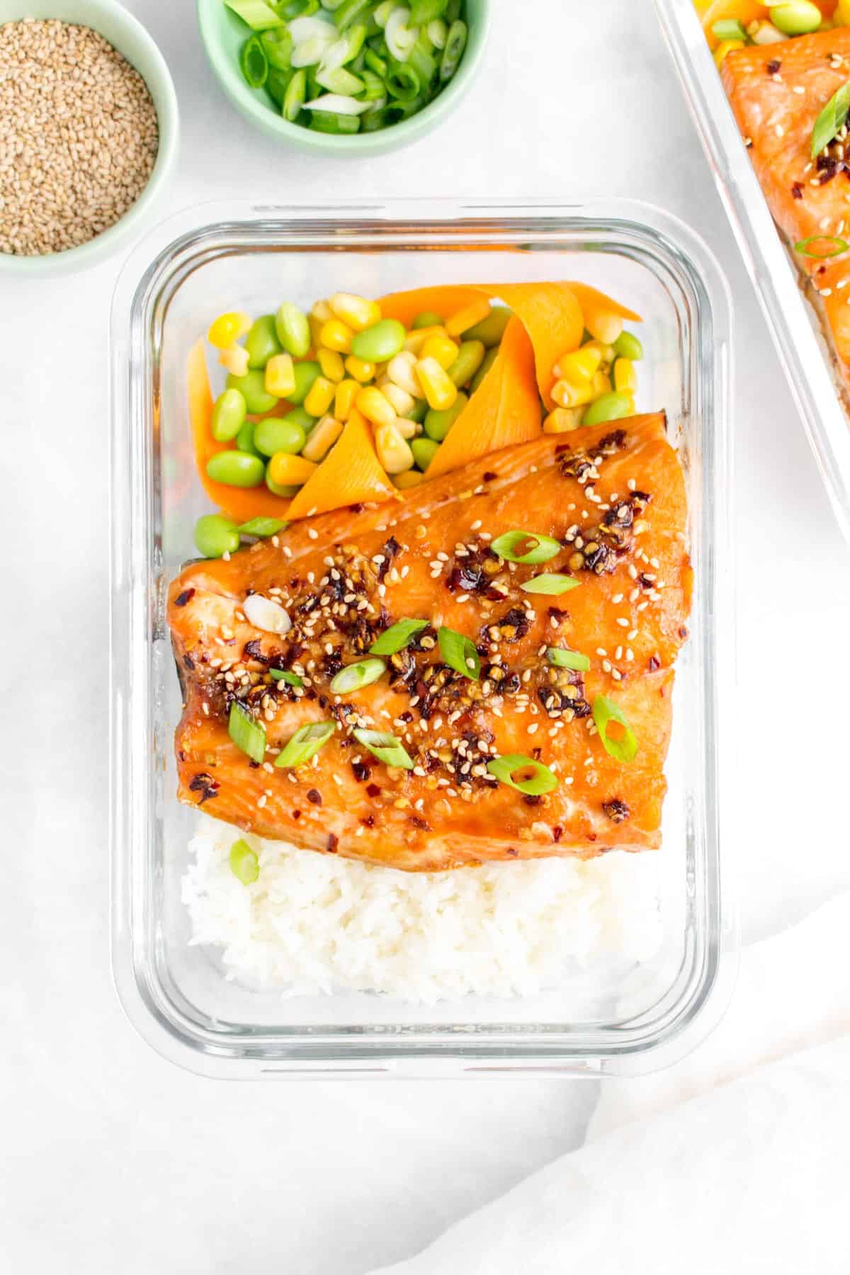 A glass meal prep container with sweet chili salmon and rice, corn, edamame, and carrots.