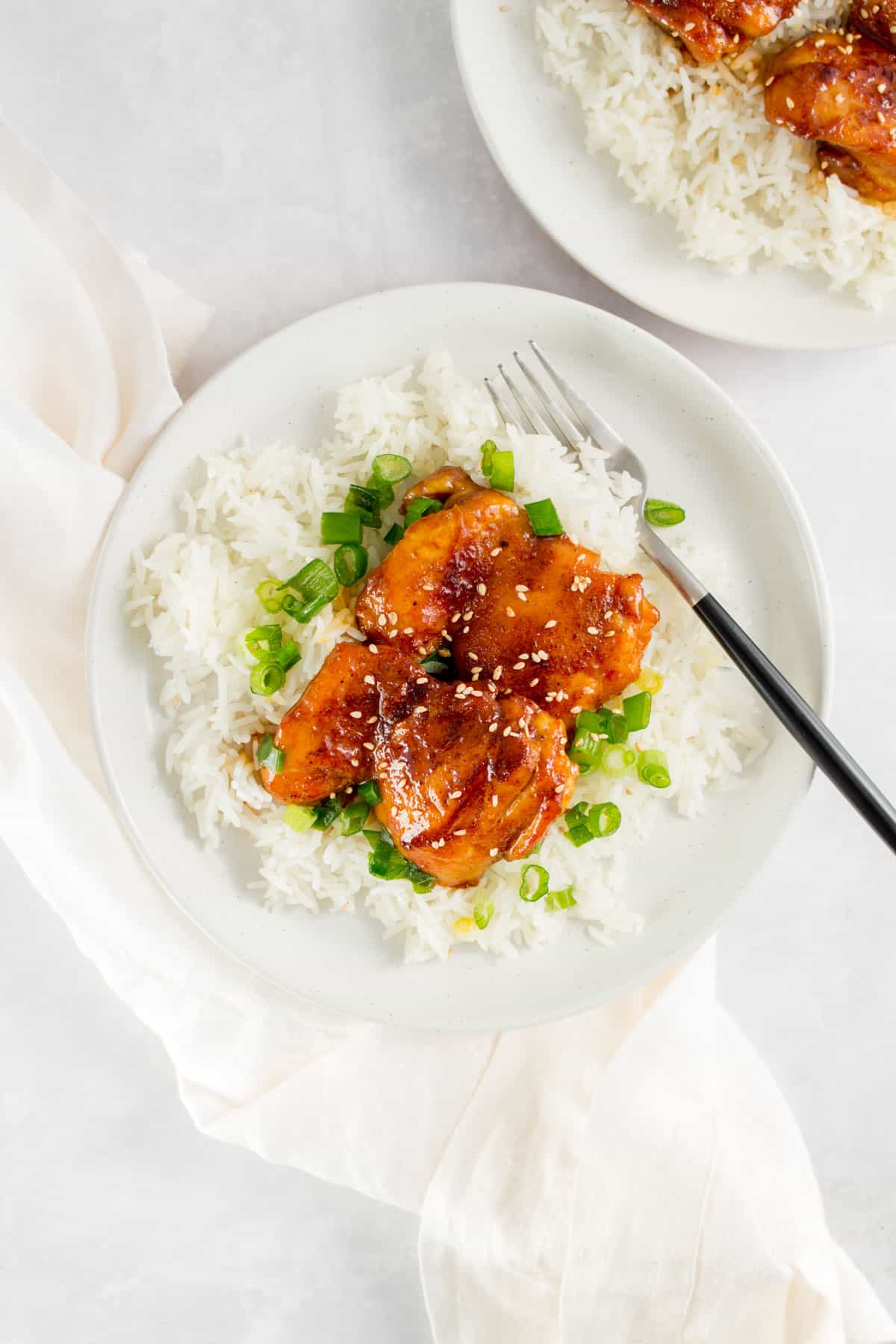 A plate with a bed of rice with green onions with two orange glazed chicken thighs on top with a fork.