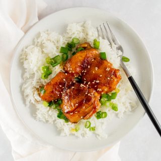 A plate with a bed of rice with green onions with two orange glazed chicken thighs on top.