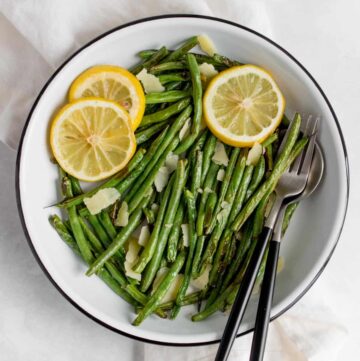 A plate of green beans with lemon slices and parmesan with a fork and a spoon inside the plate.