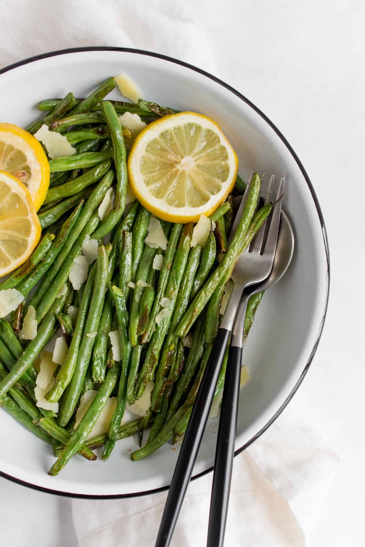 Close up of a plate of green beans with lemon slices and parmesan with a fork and a spoon inside the plate.