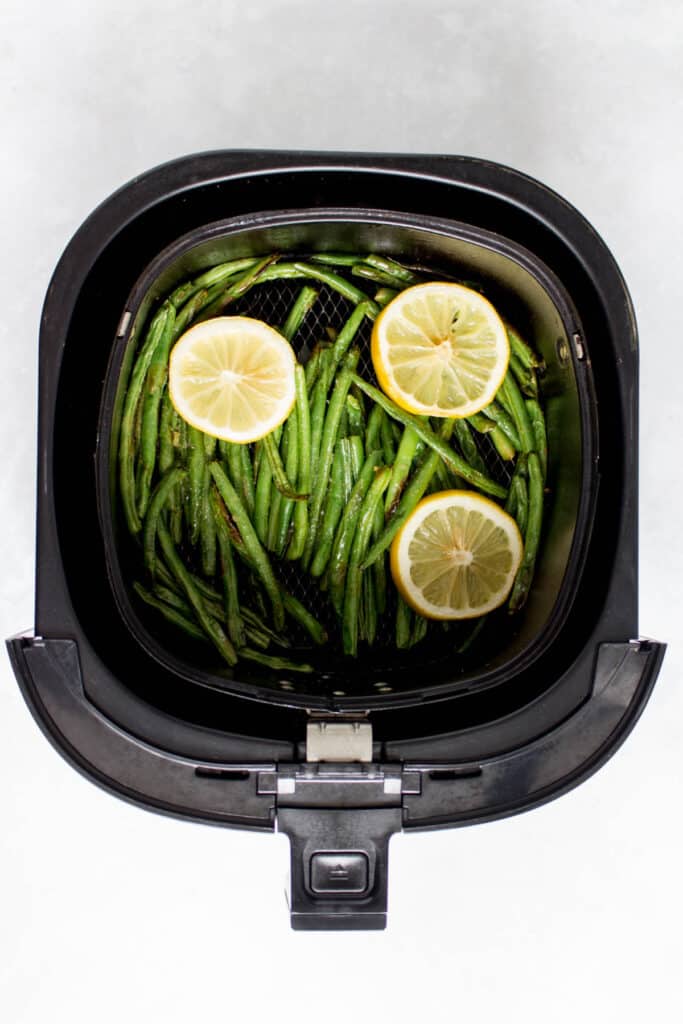 Air fryer basket with green beans with lemon slices.