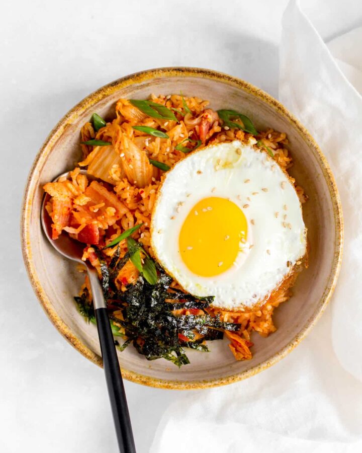 Overhead view of a bowl of bacon kimchi fried rice with a fried egg and shredded seaweed on top with a spoon tucked into the rice.