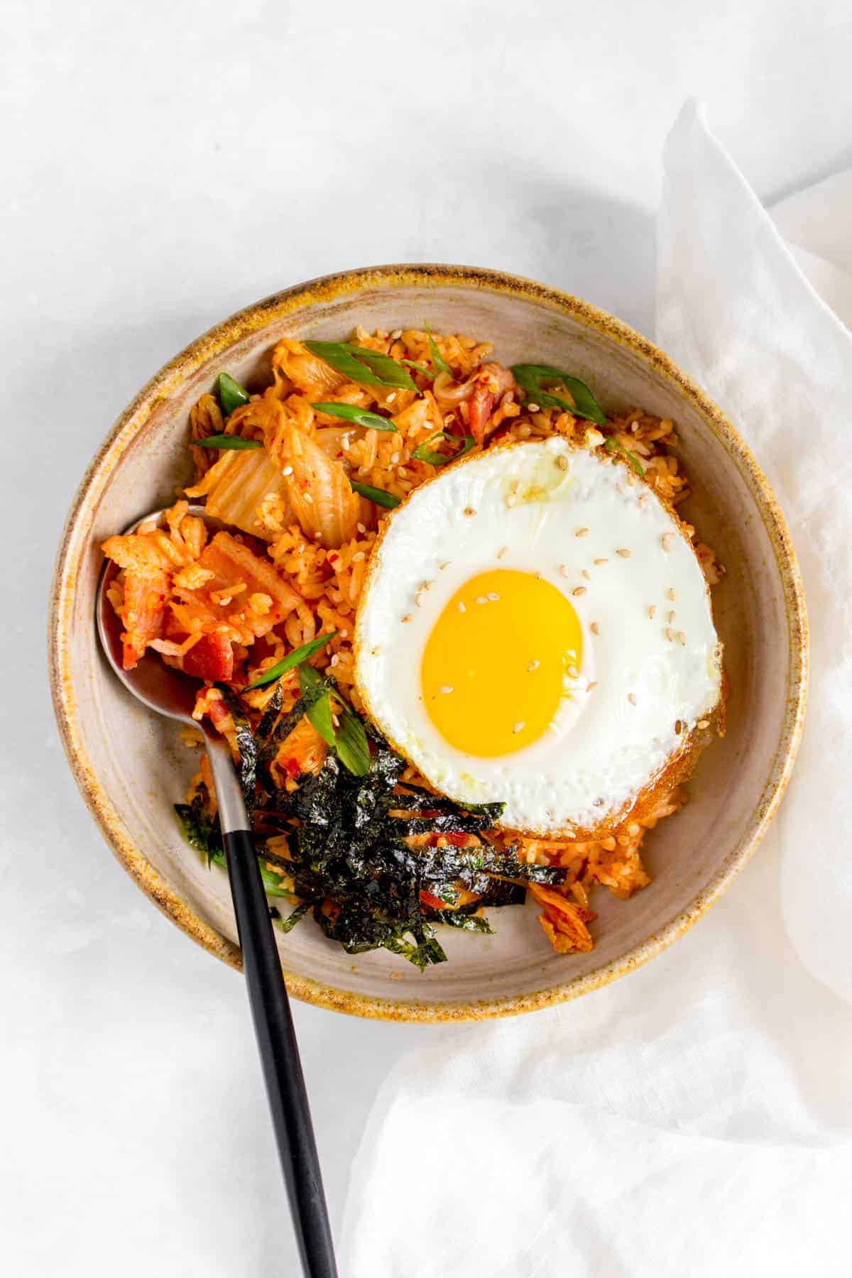 Overhead view of a bowl of bacon kimchi fried rice with a fried egg and shredded seaweed on top with a spoon tucked into the rice.