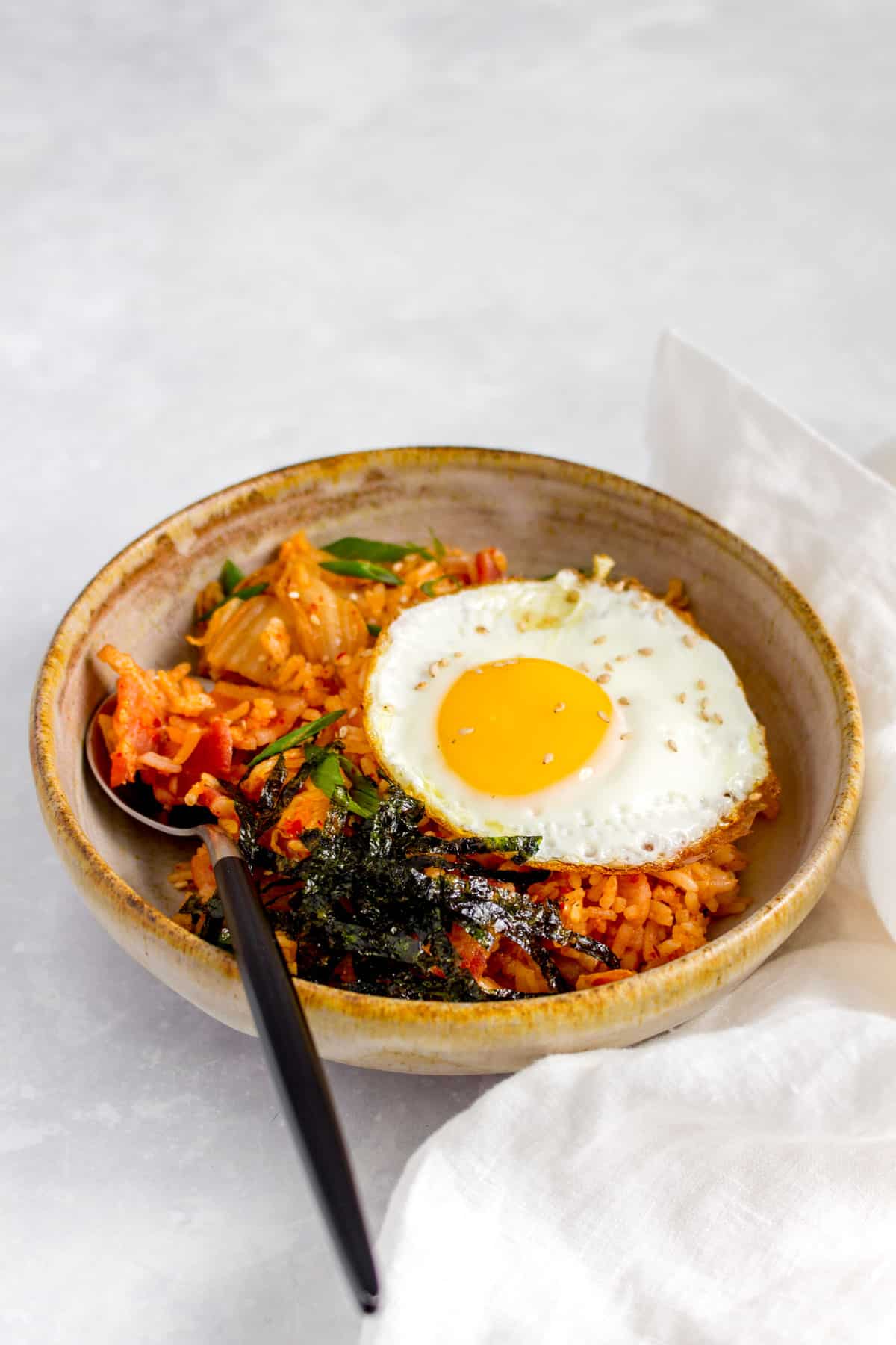 A bowl of bacon kimchi fried rice with a fried egg and shredded seaweed on top with a spoon tucked into the rice.