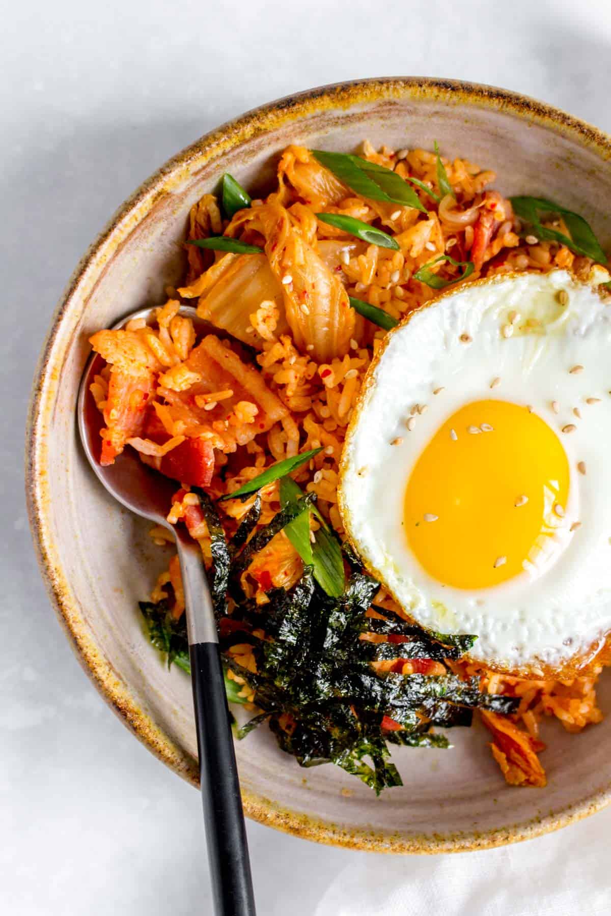 Close up of a bowl of bacon kimchi fried rice with a fried egg and shredded seaweed on top with a spoon tucked into the rice.