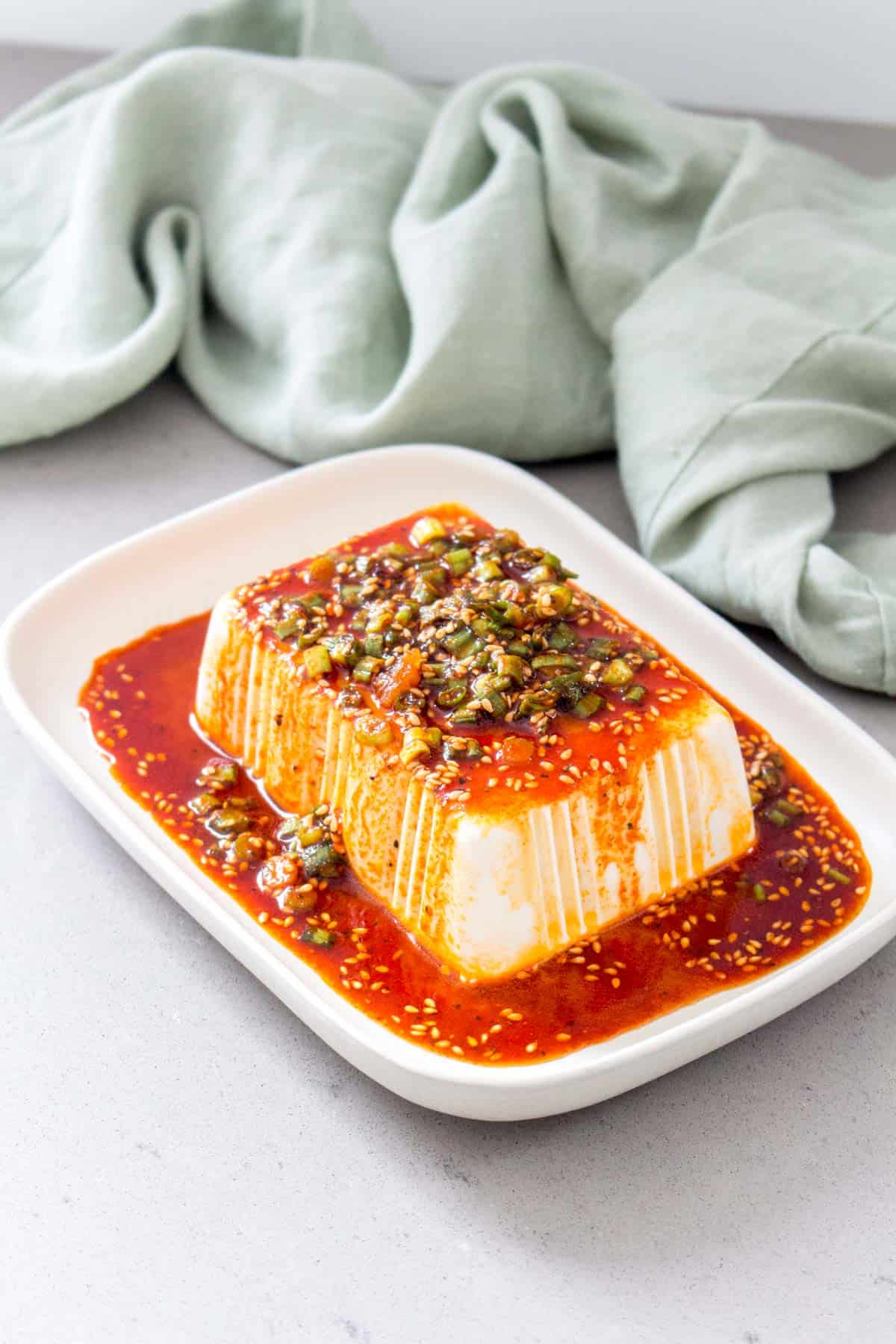 Angled view of a plate of cold silken tofu with soy sesame sauce on top and a green linen napkin in the background.