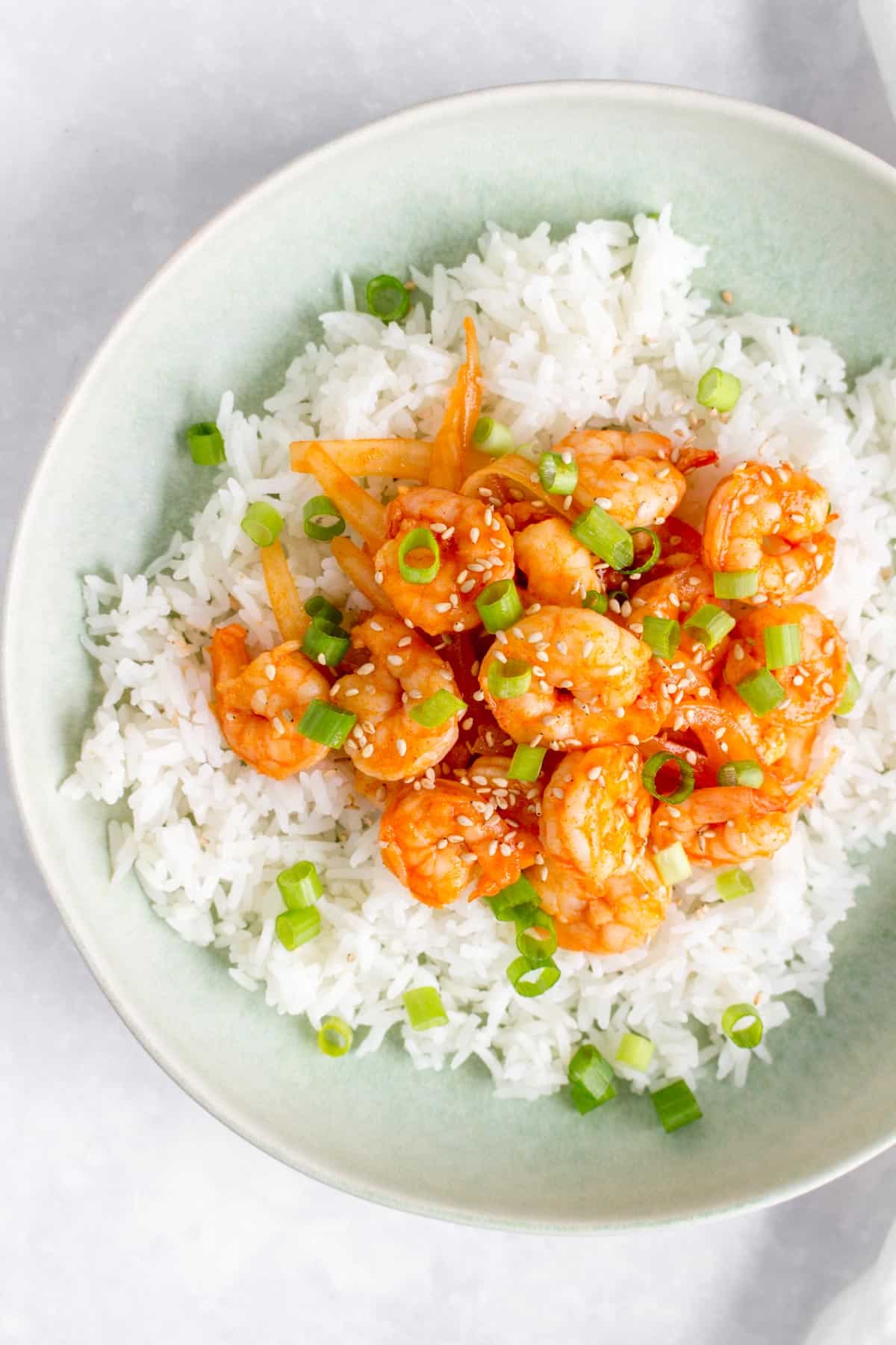 Close up of a plate of rice with sweet and spicy shrimp on top with sesame seeds and green onions.