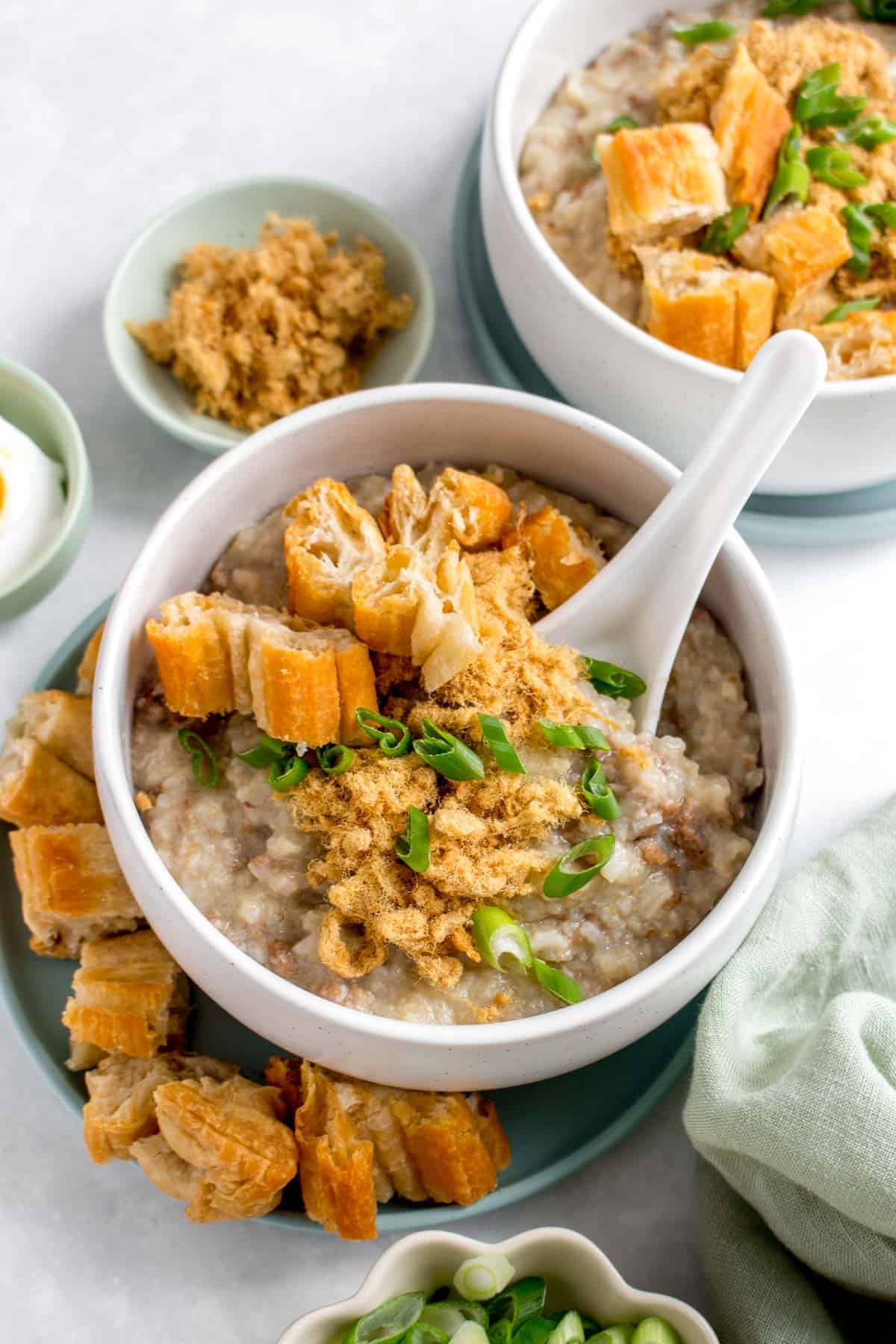 A bowl of beef congee with fried dough, pork floss, and green onions with a spoon inside.