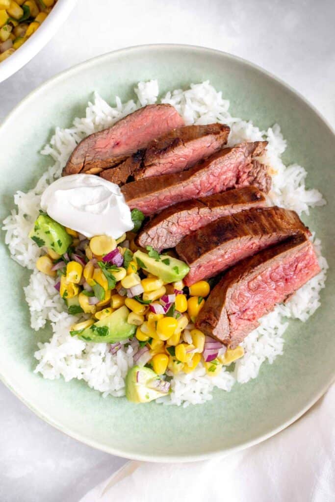 A bowl of rice with sliced flank steak, corn salsa, and sour cream.