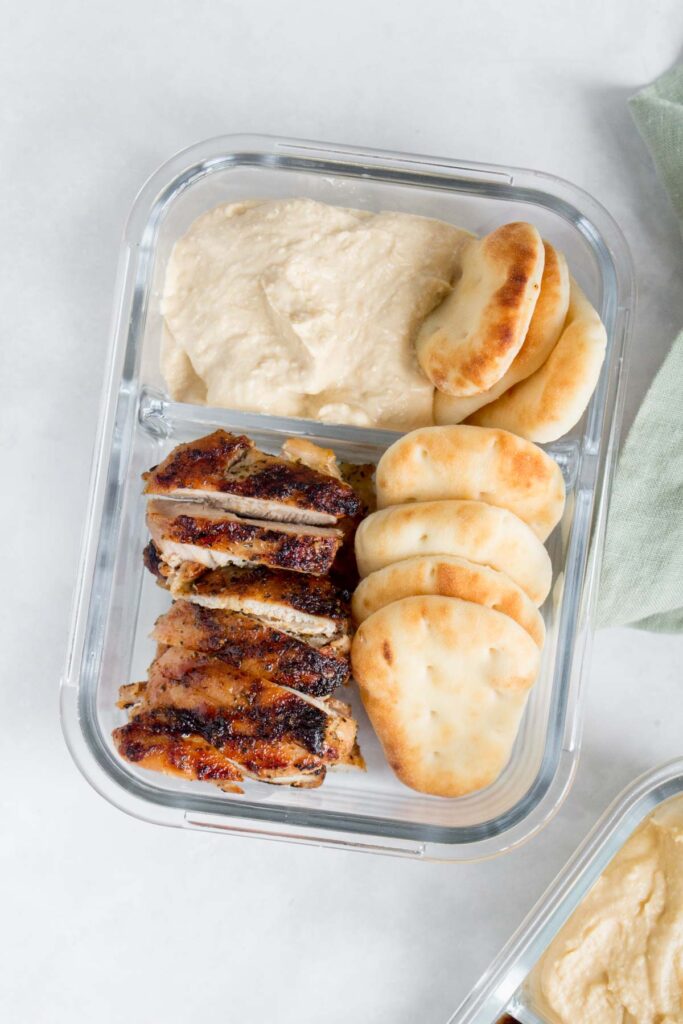 A meal prep container with two compartments, one with hummus and mini pitas/naans, and one with sliced chicken thighs and more naans/pitas.