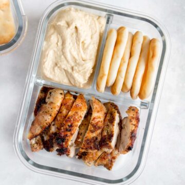 A meal prep container with three compartments, one with hummus, one with mini pitas/naans, and one with sliced chicken thighs.