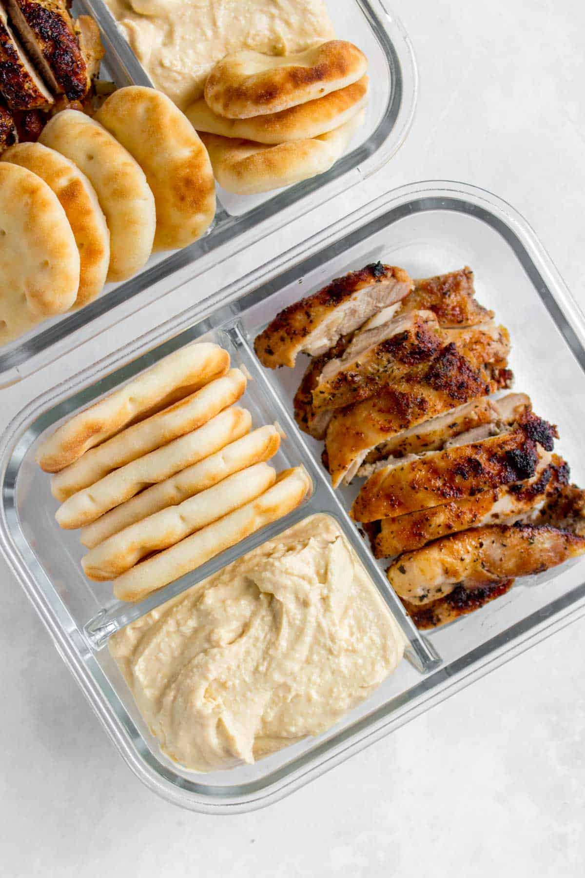 A meal prep container with three compartments, one with hummus, one with mini pitas/naans, and one with sliced chicken thighs.