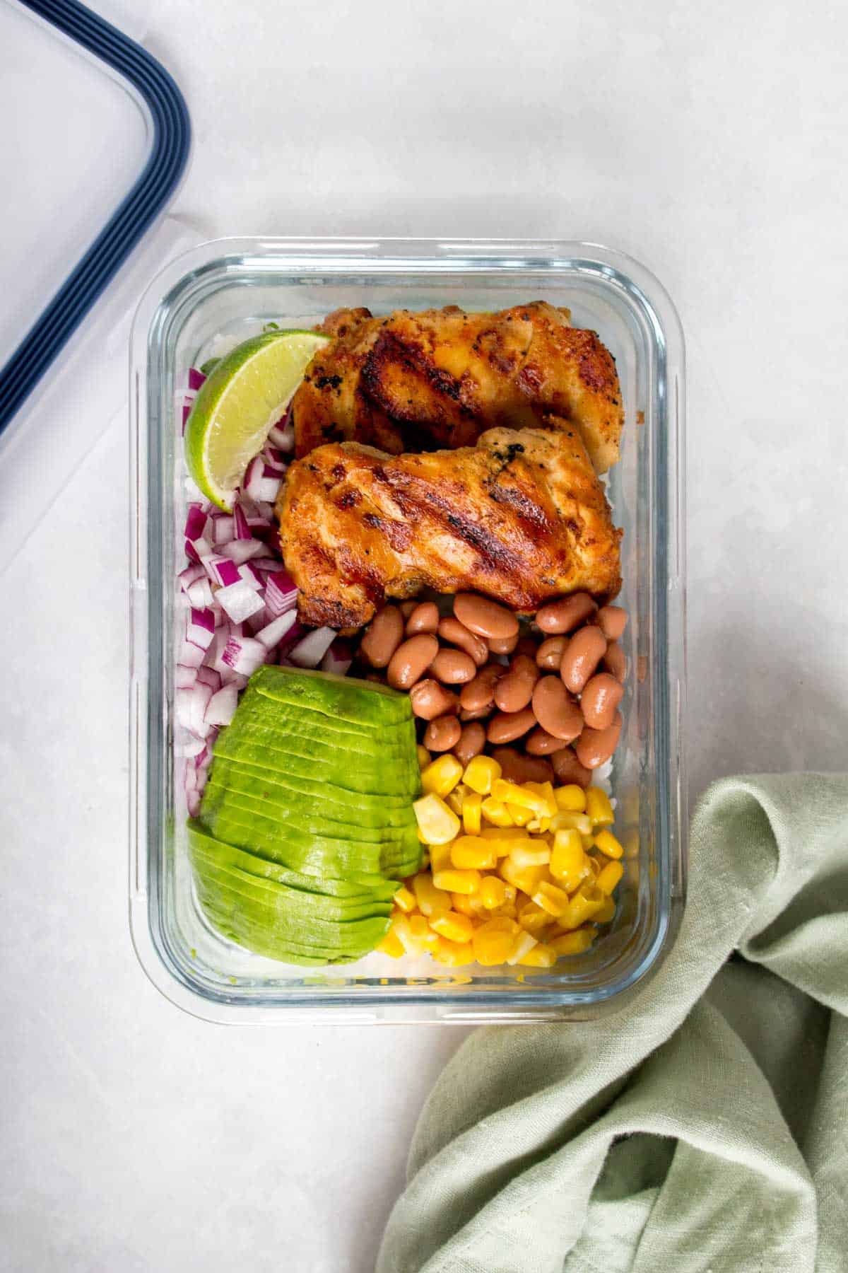 Meal prep container with grilled cilantro lime chicken thighs, corn, pinto beans, red onions, sliced avocado, and a wedge of lime.