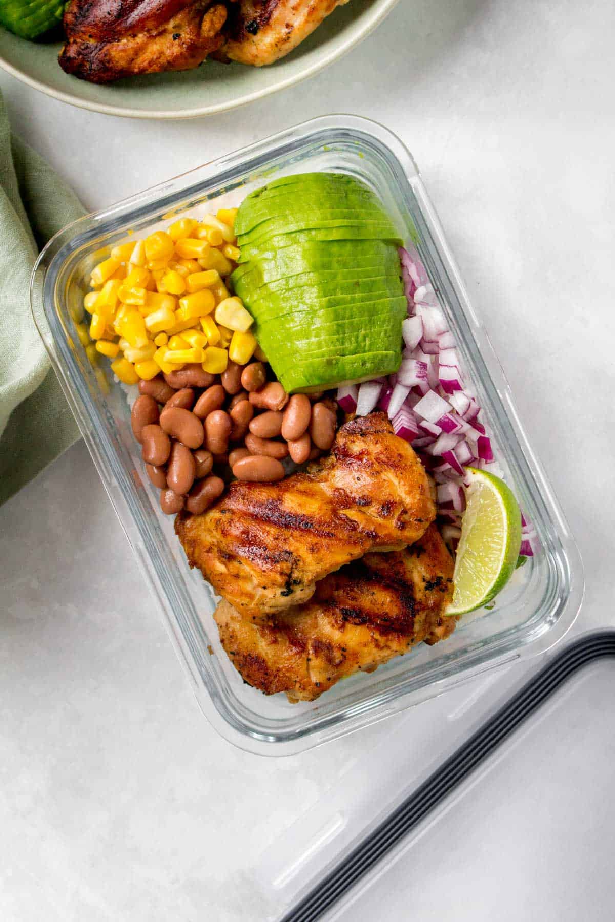 Meal prep container with grilled cilantro lime chicken thighs, corn, pinto beans, red onions, sliced avocado, and a wedge of lime.