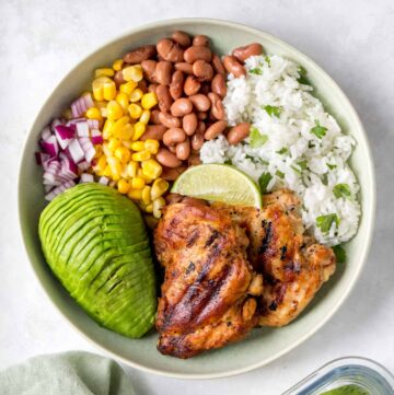 A green plate with grilled cilantro lime chicken thighs, cilantro lime rice, corn, pinto beans, red onions, sliced avocado, and a wedge of lime.