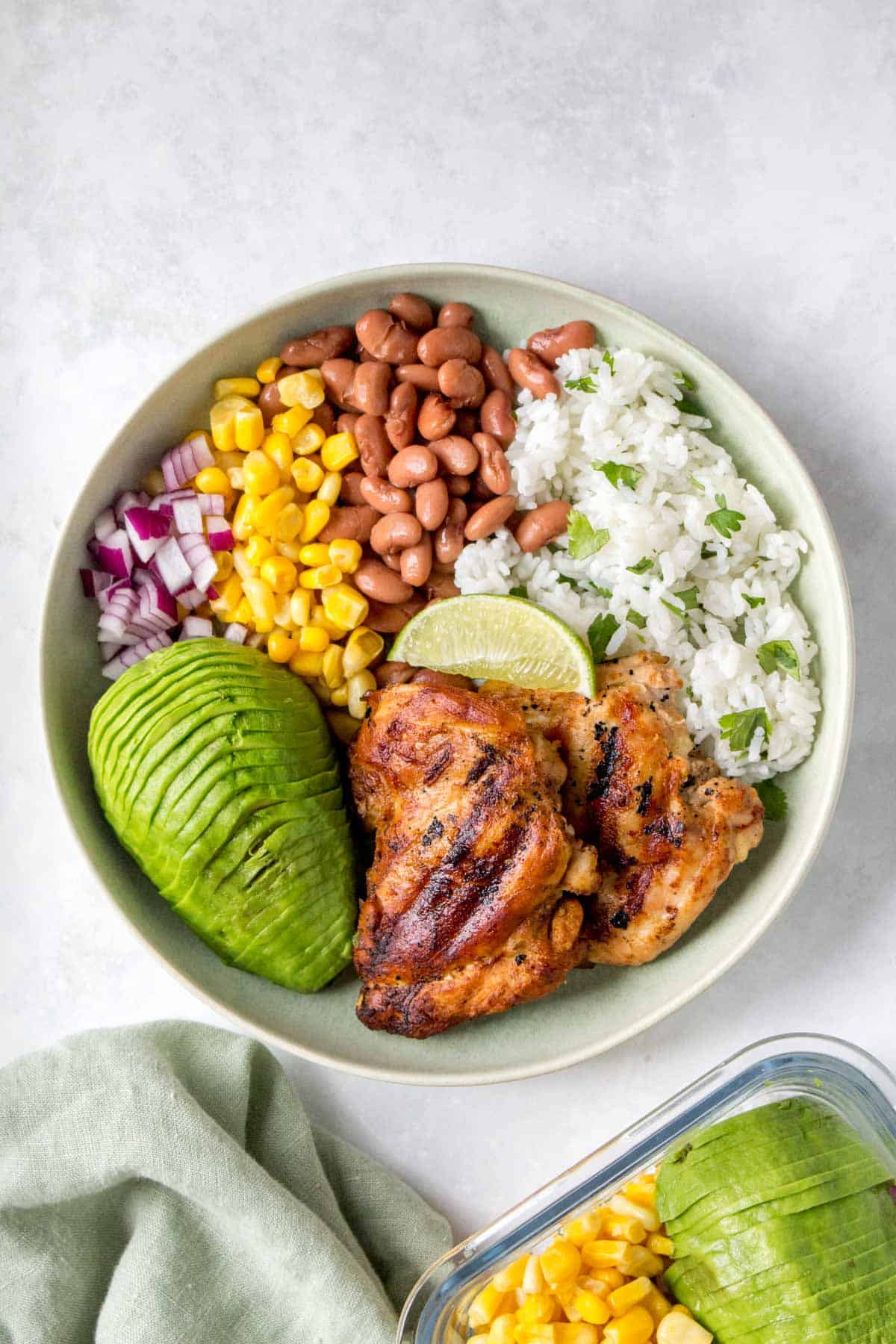 A green plate with grilled cilantro lime chicken thighs, cilantro lime rice, corn, pinto beans, red onions, sliced avocado, and a wedge of lime.