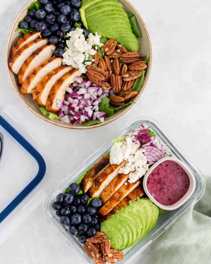 A bowl and a meal prep container with grilled chicken salad with blueberries, pecans, onions, avocado, and goat cheese with a blueberry vinaigrette.