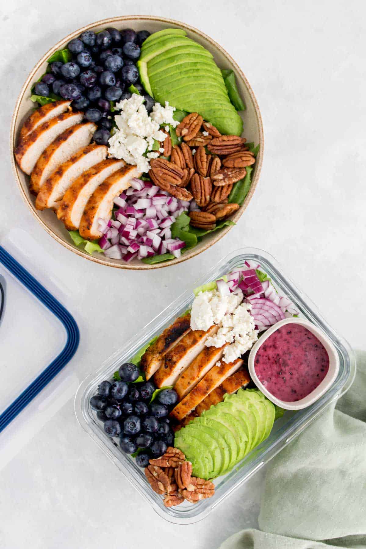 A bowl and a meal prep container with grilled chicken salad with blueberries, pecans, onions, avocado, and goat cheese with a blueberry vinaigrette.