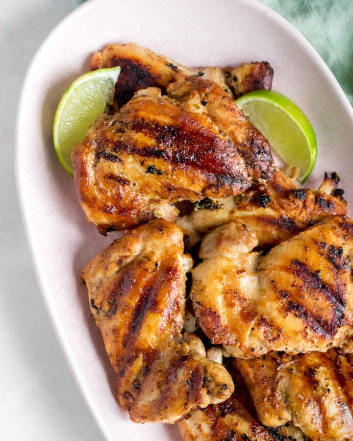 Close up of a pink platter of grilled cilantro lime chicken thighs with lime wedges.