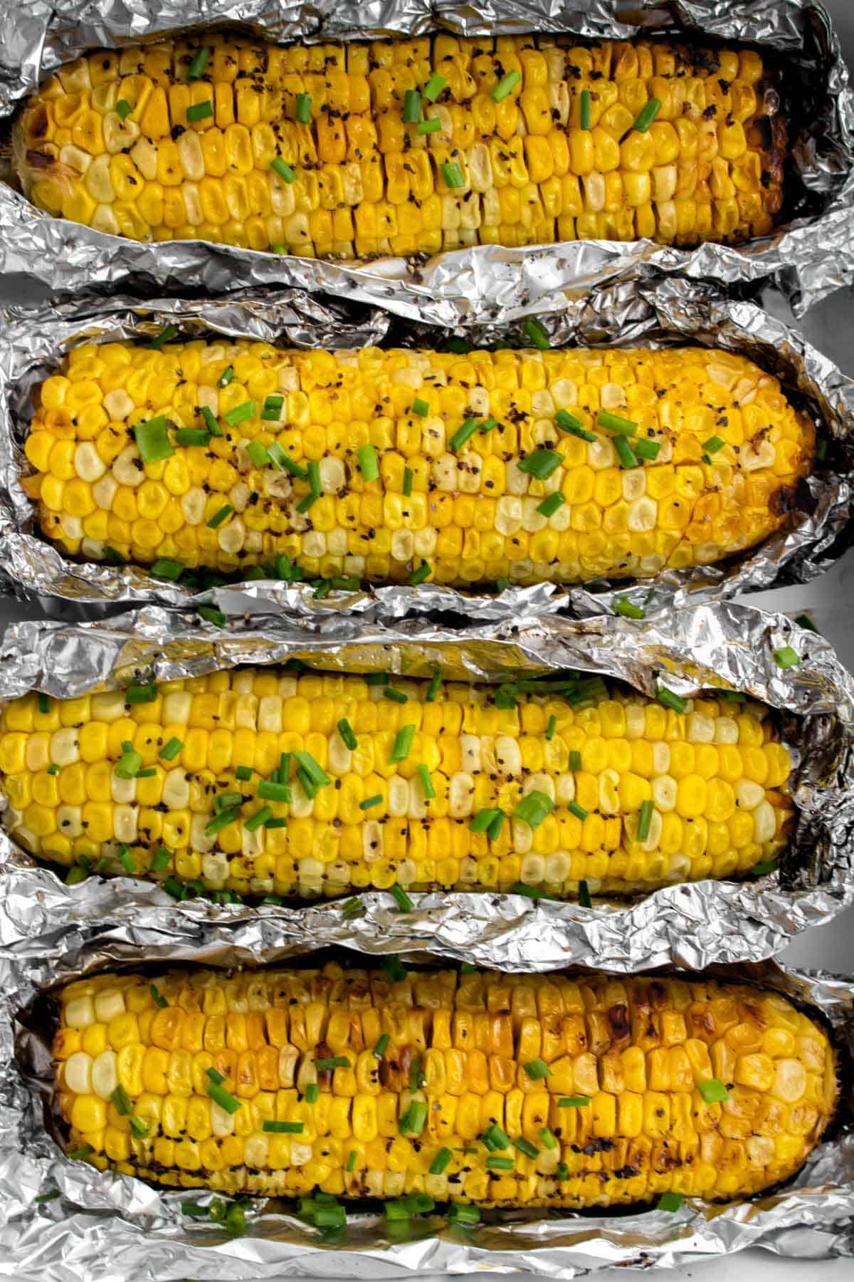 Overhead view of four corn on the cob with tin foil around them with chopped chives on top.