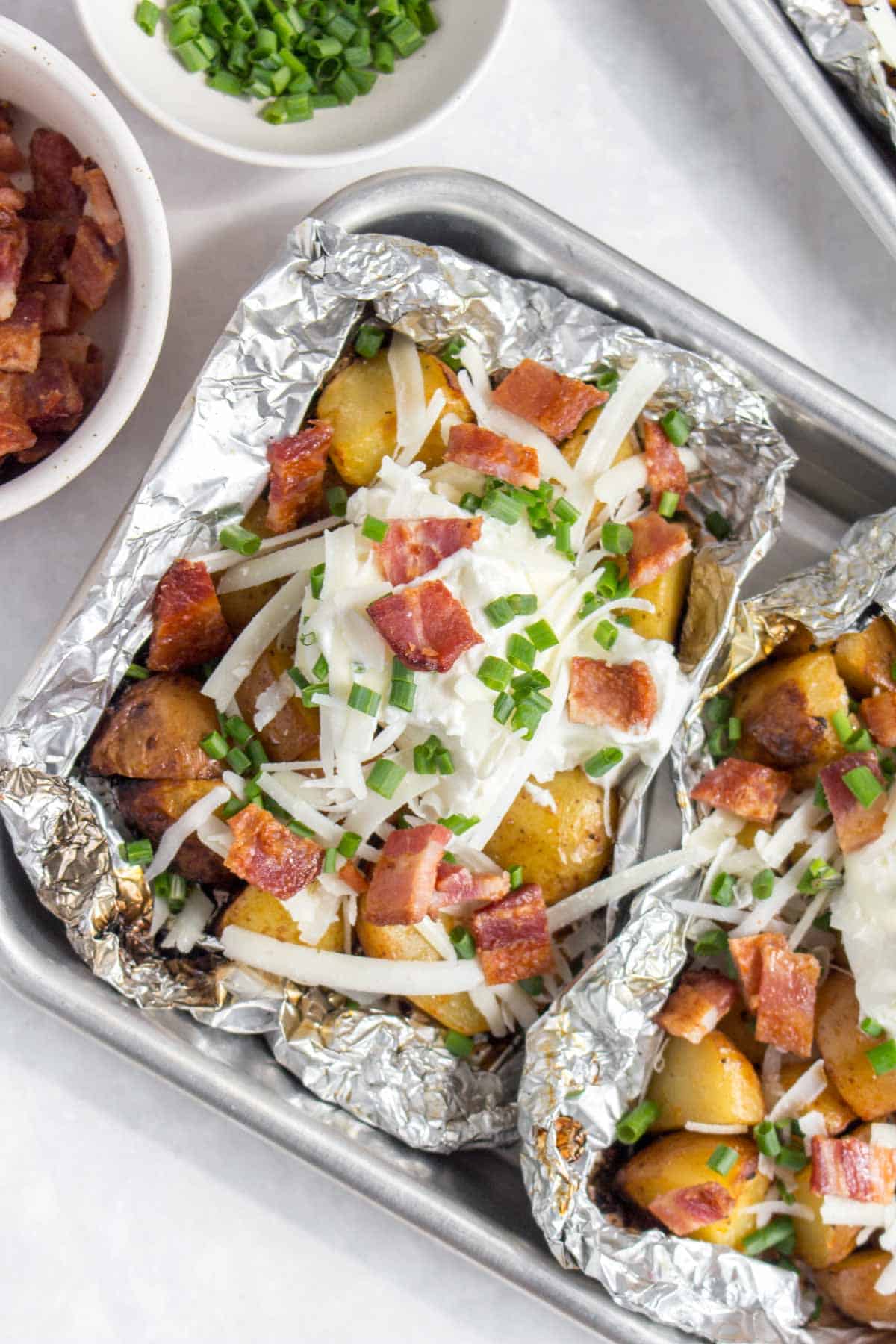 Overhead view of two foil packet potatoes with sour cream, cheese, bacon, and chives.
