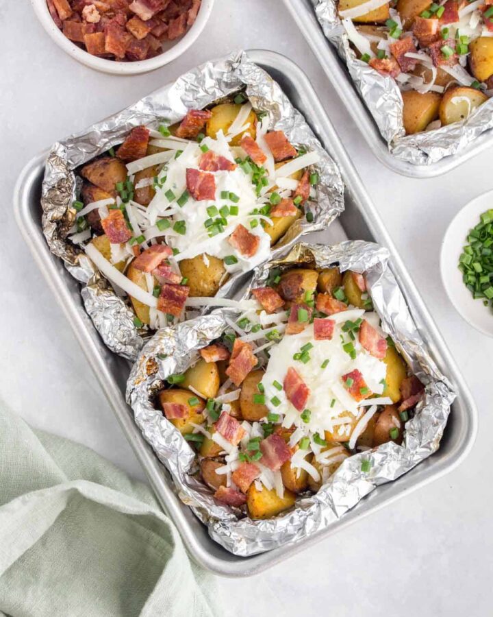 Overhead view of two foil packet potatoes on a sheet pan with sour cream, cheese, bacon, and chives.