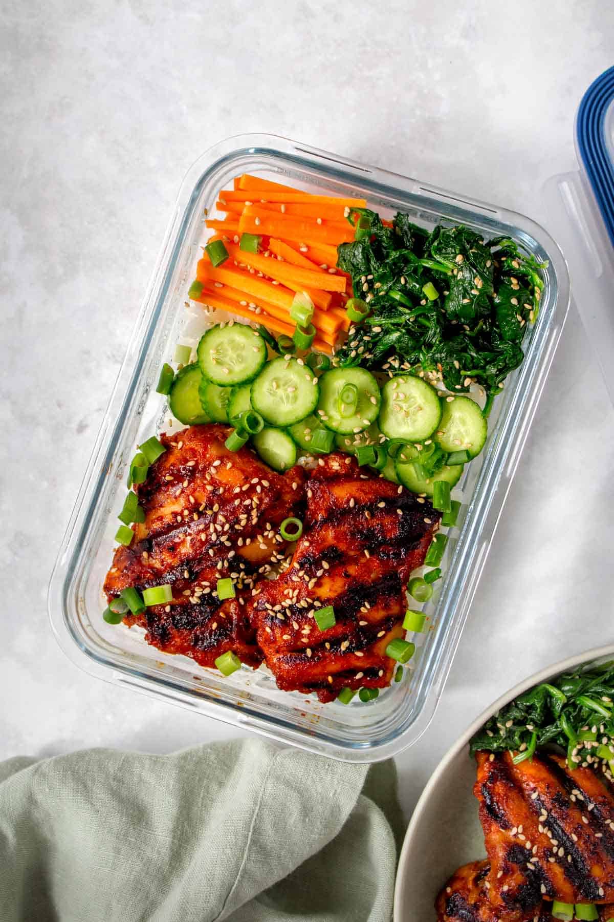 Meal prep container with rice, korean chicken thighs, cucumbers, carrots, seasoned seaweed, sesame seeds, and green onions.