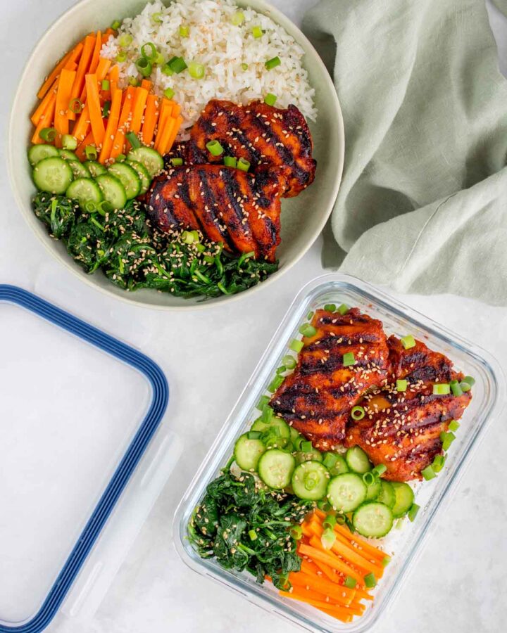Plate and meal prep container with rice, korean chicken thighs, cucumbers, carrots, seasoned seaweed, sesame seeds, and green onions.