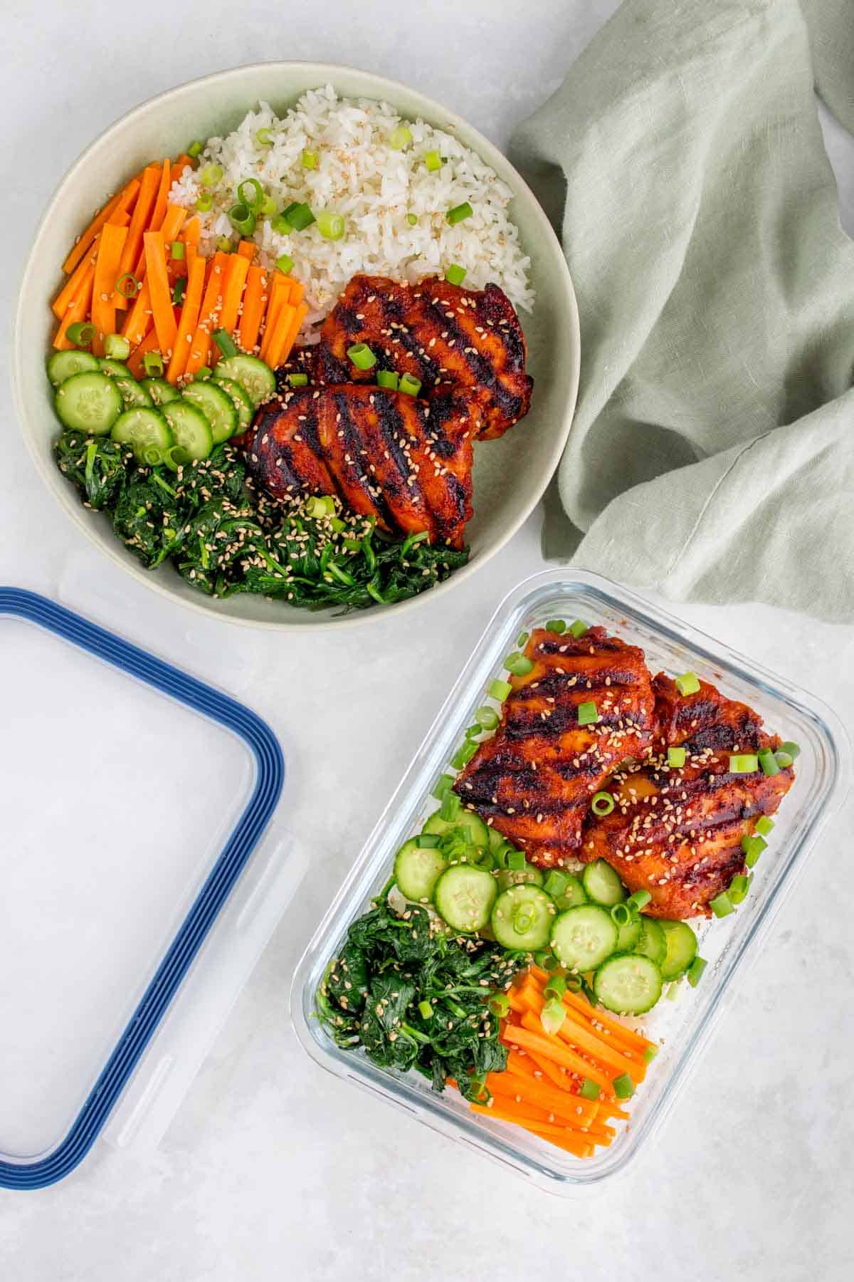 Plate and meal prep container with rice, korean chicken thighs, cucumbers, carrots, seasoned seaweed, sesame seeds, and green onions.