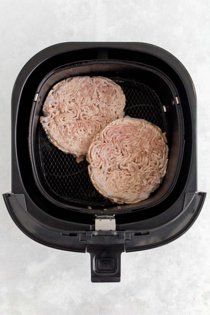 Two frozen hamburger patties in an air fryer basket.