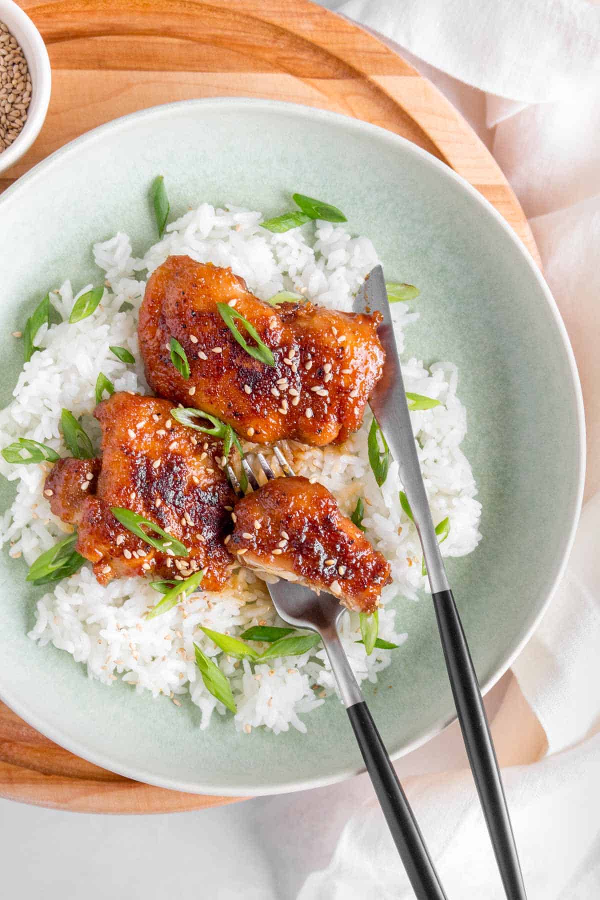 A plate with two pieces of teriyaki chicken thighs, one cut, with a fork and knife in the plate.