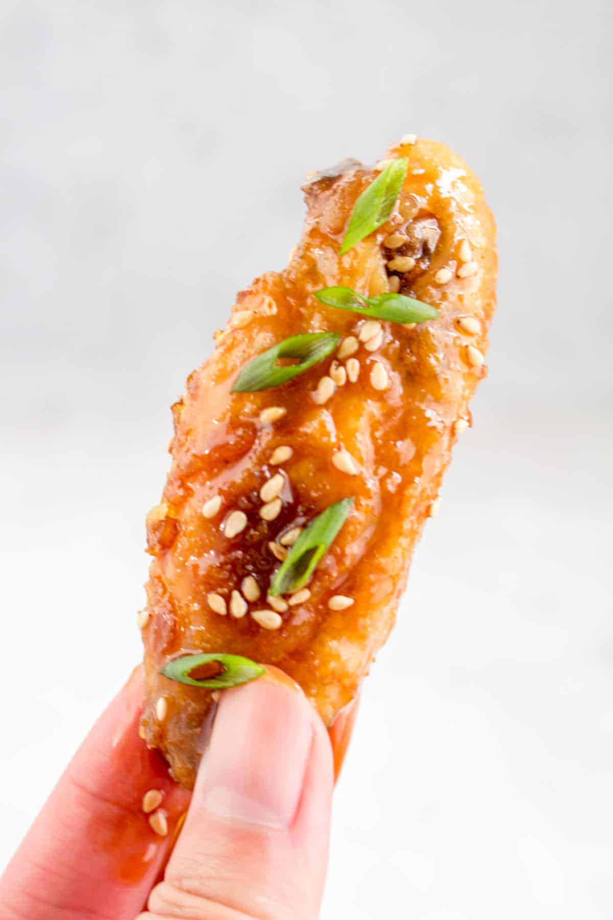 A hand holding a single chicken wing with teriyaki sauce, green onions, and sesame seeds.