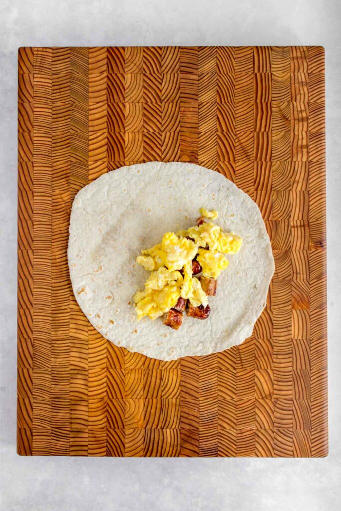 A tortilla wrap with cut sausages and scrambled eggs on a serving board.