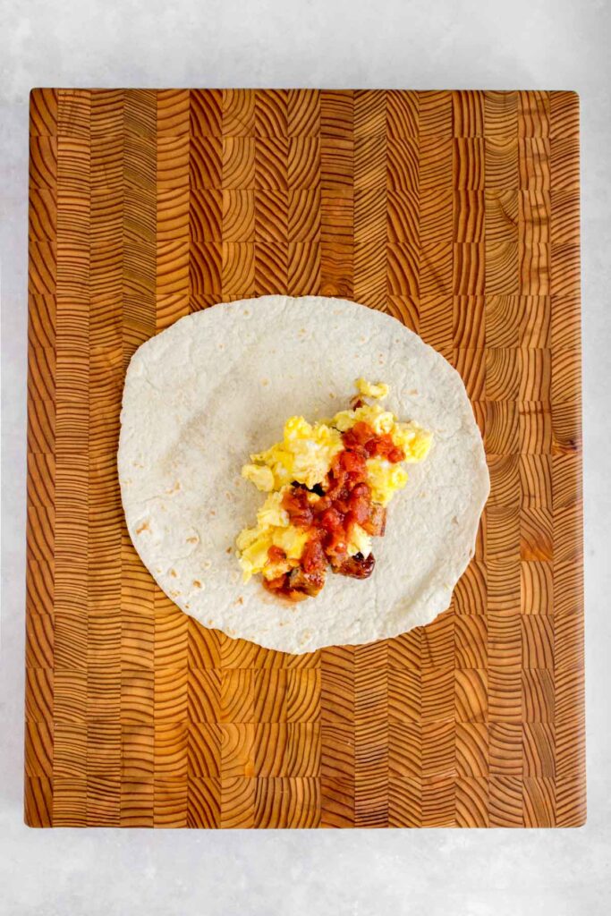 A tortilla wrap with cut sausages, scrambled eggs, and salsa on a serving board.
