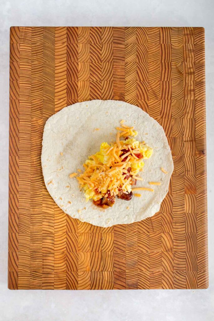 A tortilla wrap with cut sausages, scrambled eggs, and salsa on a serving board with shredded cheese on top.