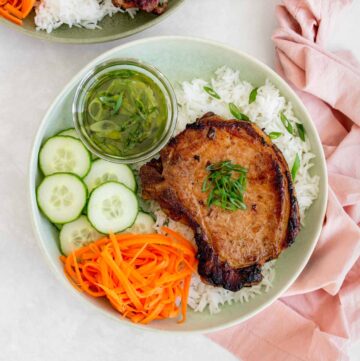A plate with Vietnamese pork chop on top of rice with scallion oil, cucumbers, and pickled carrots.