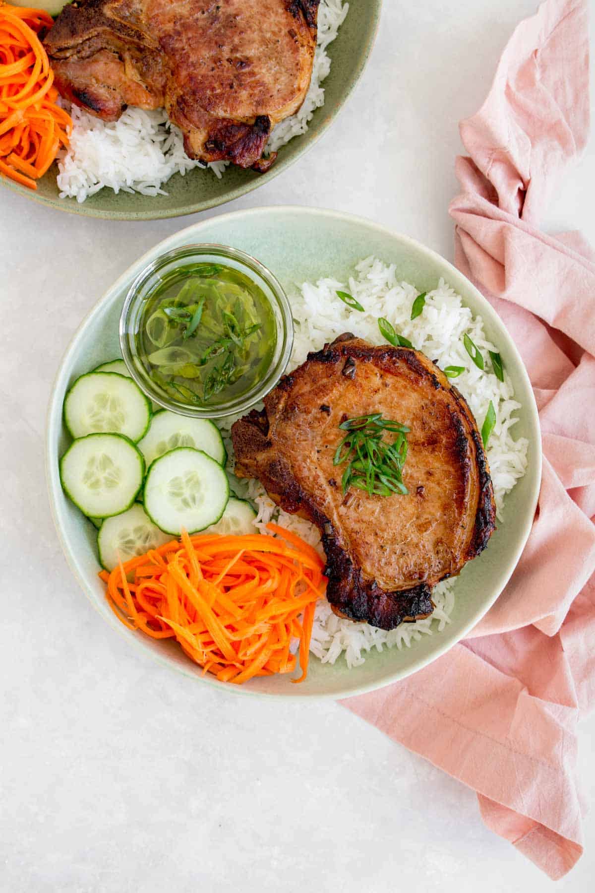 A plate with Vietnamese pork chop on top of rice with scallion oil, cucumbers, and pickled carrots.