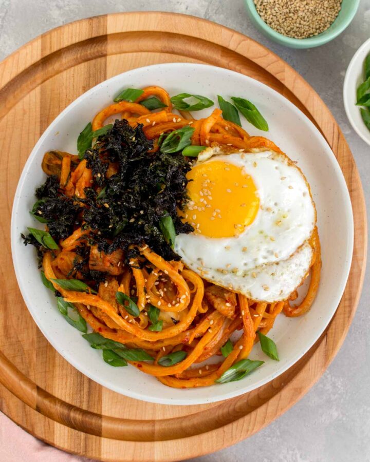 Overhead view of a plate of kimchi udon with a fried egg and seaweed on top.